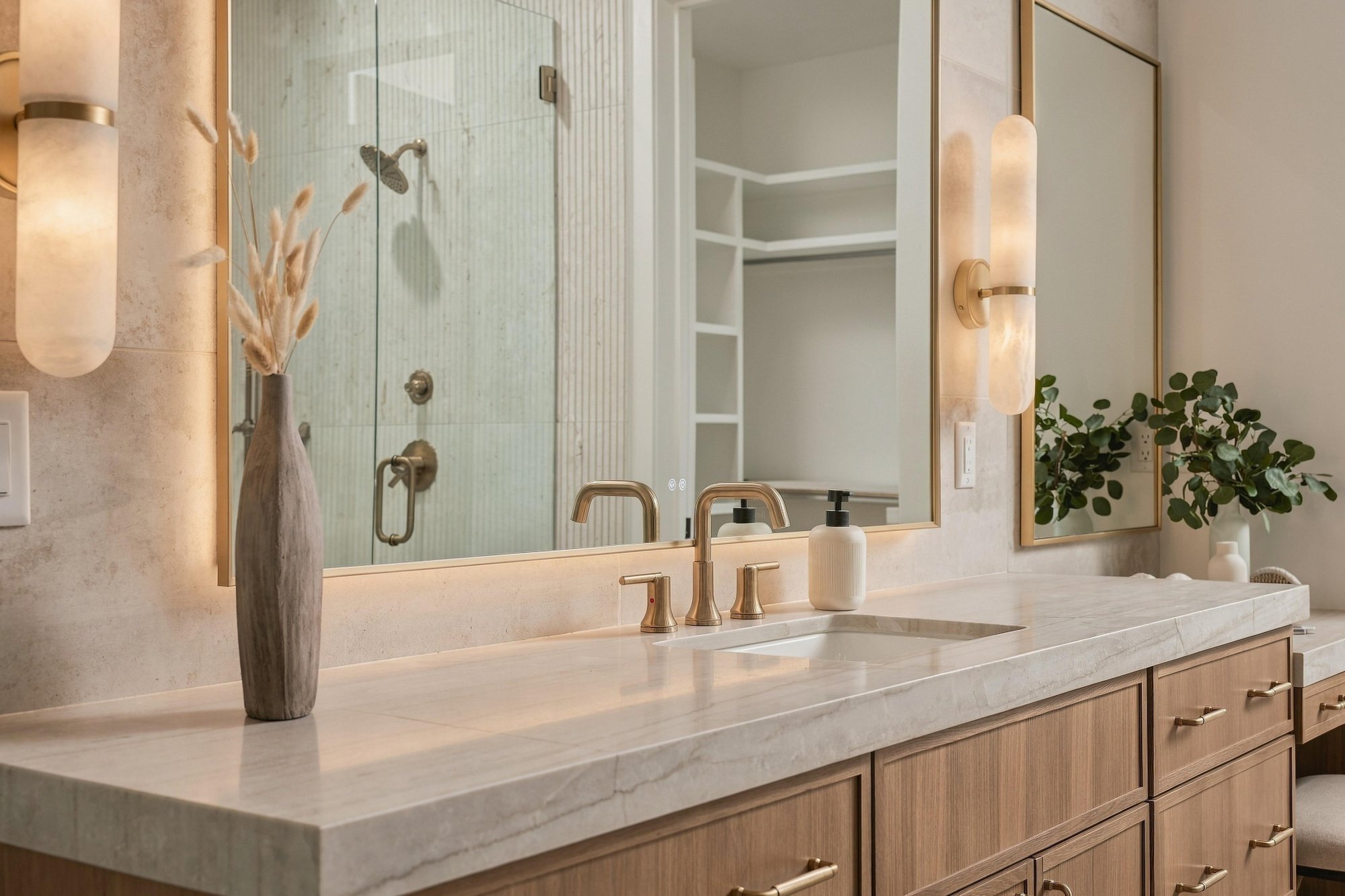 Primary vanity with brass faucets and alabaster sconces beside a fluted glass shower
